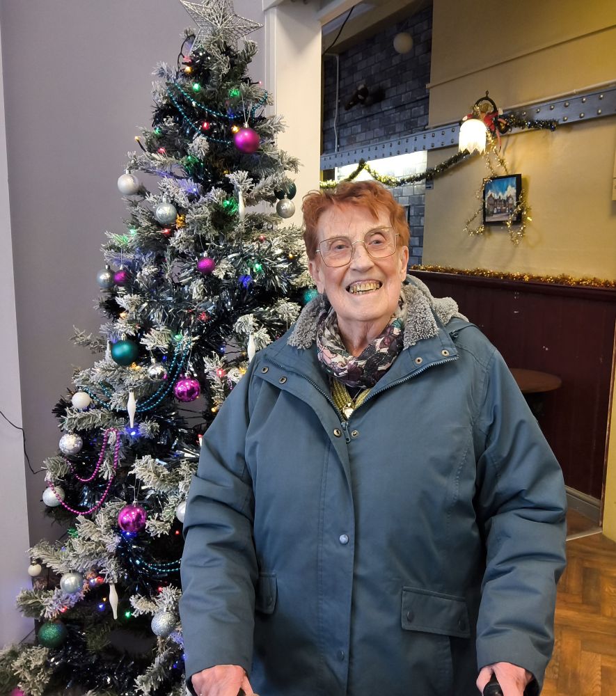 A woman grins broadly towards the camera, a Christmas tree behind her to the left