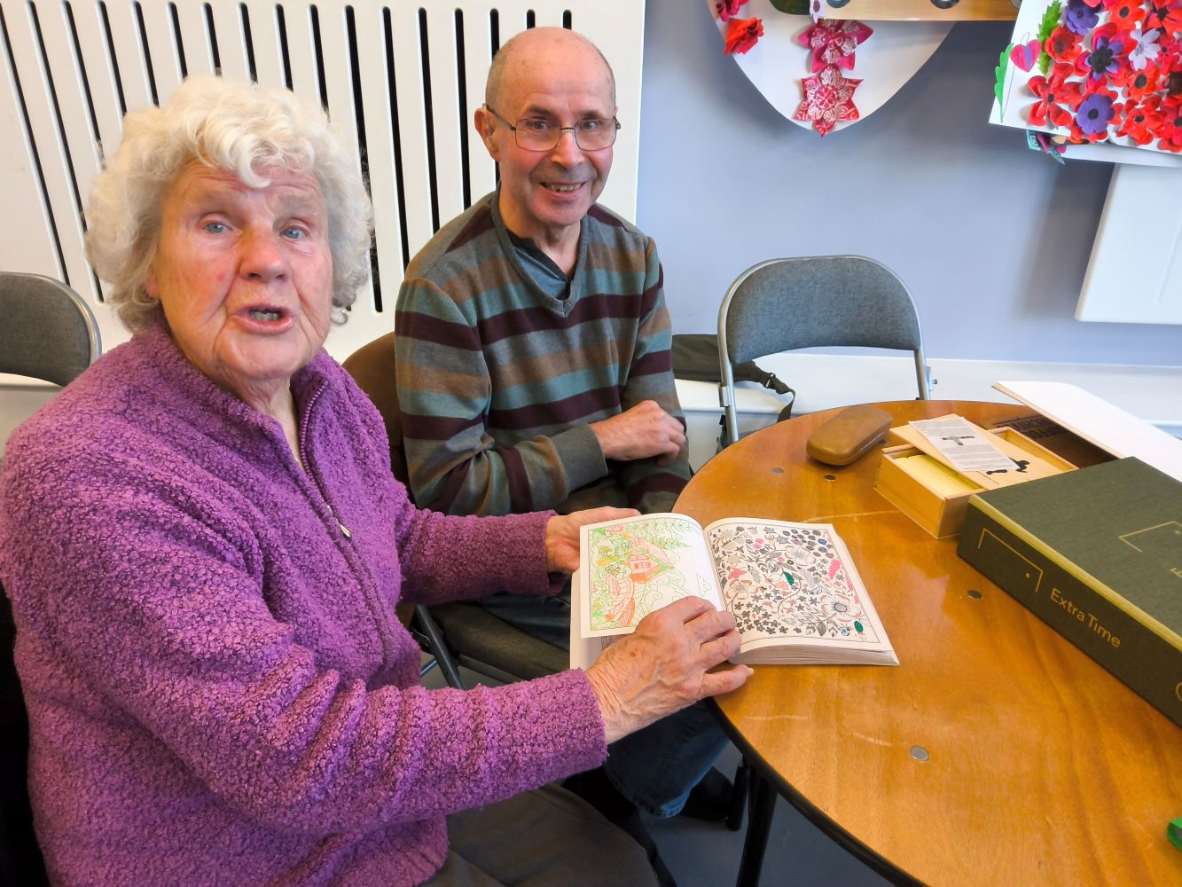 A gentleman dressed in a striped V-neck jumper and with arms crossed smiles towards the camera whilst a woman seated beside him, dressed in a purple zipped top holds a Colouring for Mindfulness book open at a page she's been working on