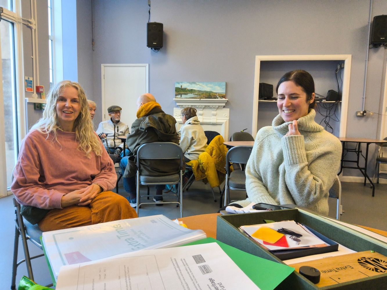 Two women sit a table strewn with papers and the Extra Time Story Box. They are smiling as one holds the large collar of her woolly jumper and the other, hands crossed in her lap, looks towards the camera