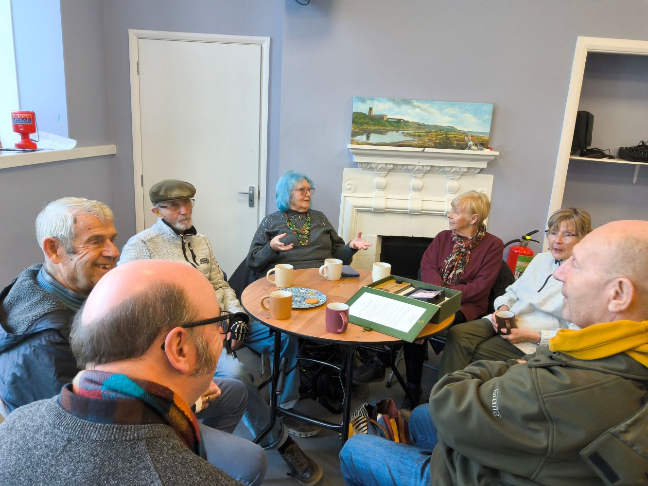 A group of adults seated around a table. In the background, right, is a white door beside which is a firesurround with a landscape painting on the top and shelves to the other side. On the table are cups and a plate with a single biscuit on it. Some people appear to be talking in an animated way, whilst others sit looking on, arms crossed