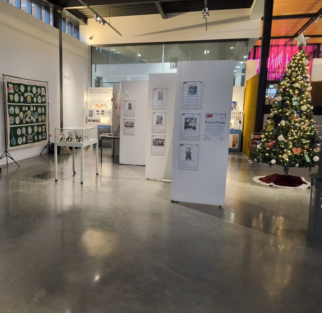 It is an exhibition space, with large white panels and a banner displayed against a white wall. To the right is a Christmas tree and there is a glass cabinet on legs with objects inside
