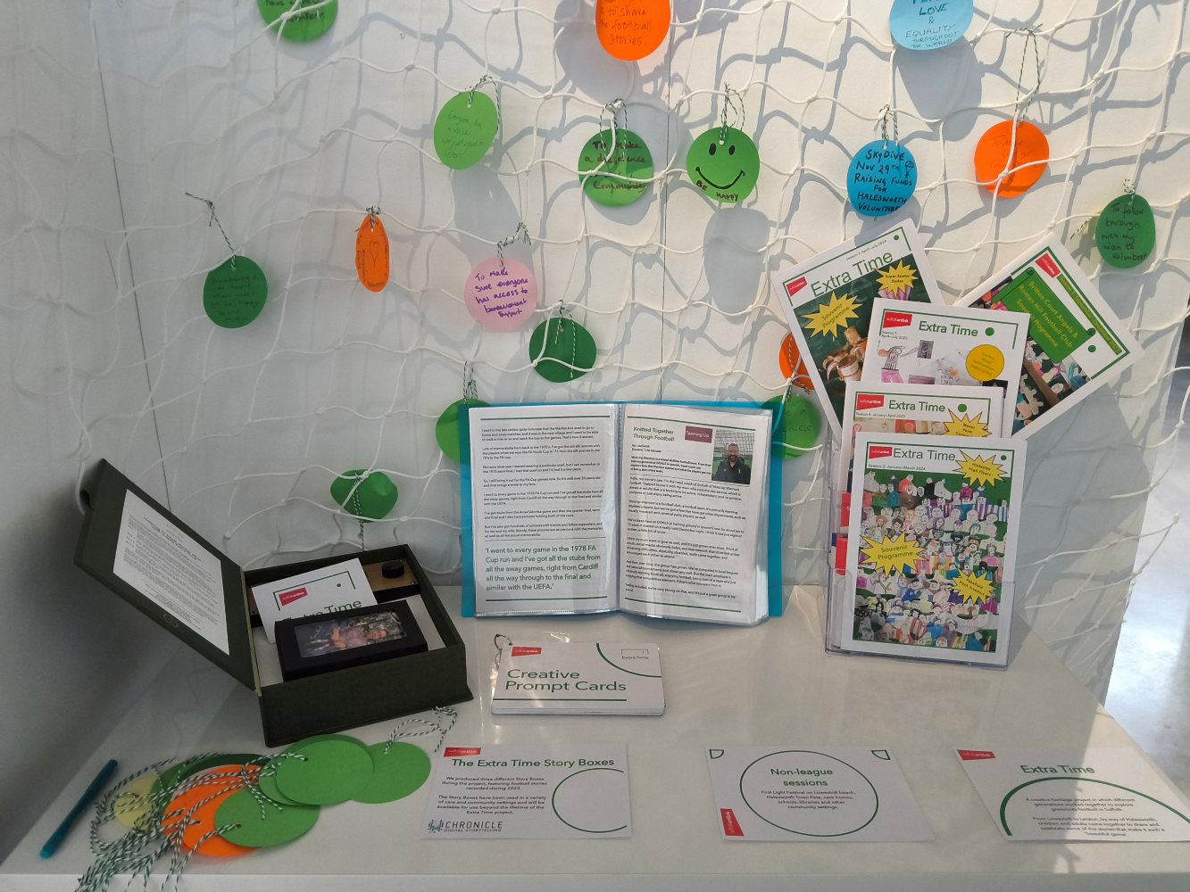 A table set against a white panel on which is draped a football net with several coloured discs tied to it. On the table is a small stand holding A4 Souvenir Programmes, a booklet opened at a story about football and a story box, lid open, to the left. There's also a pile of green and orange discs.