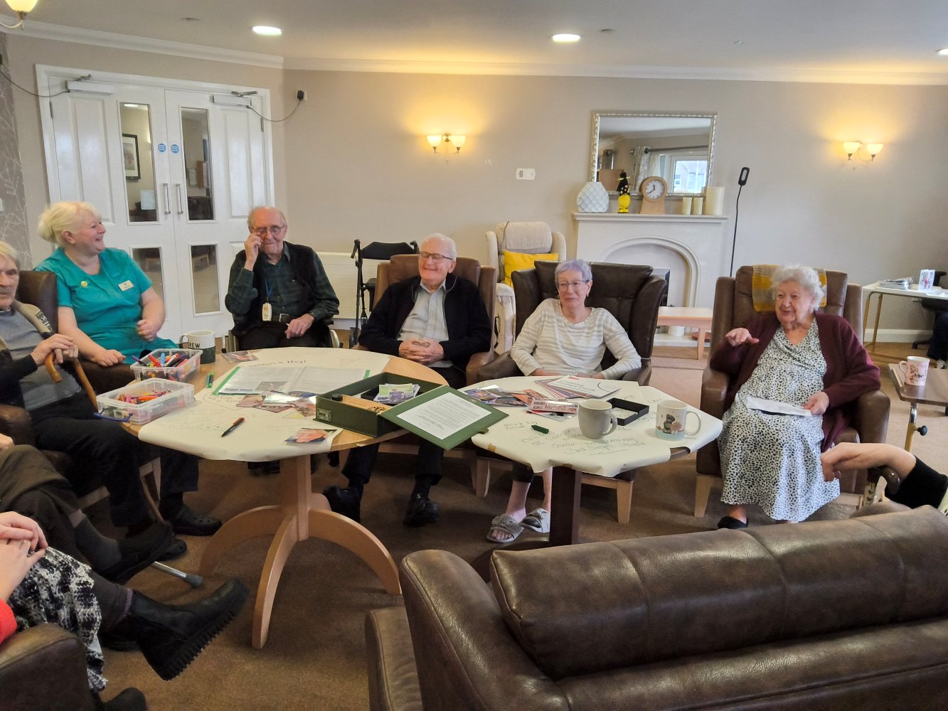 A group of residents and the care staff having a chat, whilst in front of them on two circular tables is a collection of photographs, mugs and other items