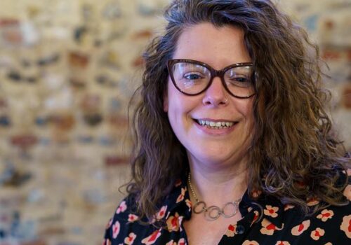 A white woman with brown framed glasses and brown curly hair is standing in front of a brick wall.