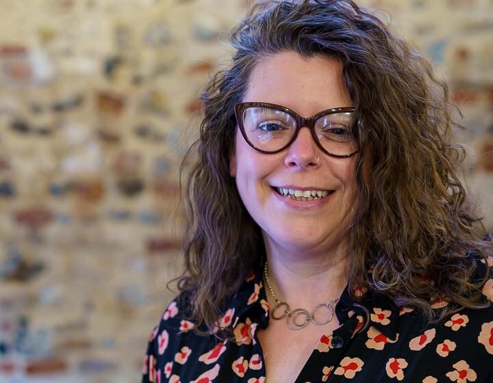 A white woman with brown framed glasses and brown curly hair is standing in front of a brick wall.