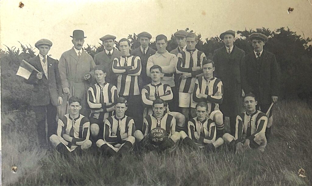A monochrome photograph of a football team, in the centre a player holding the football with the words Boiler Shop 1917 - 1918 on it