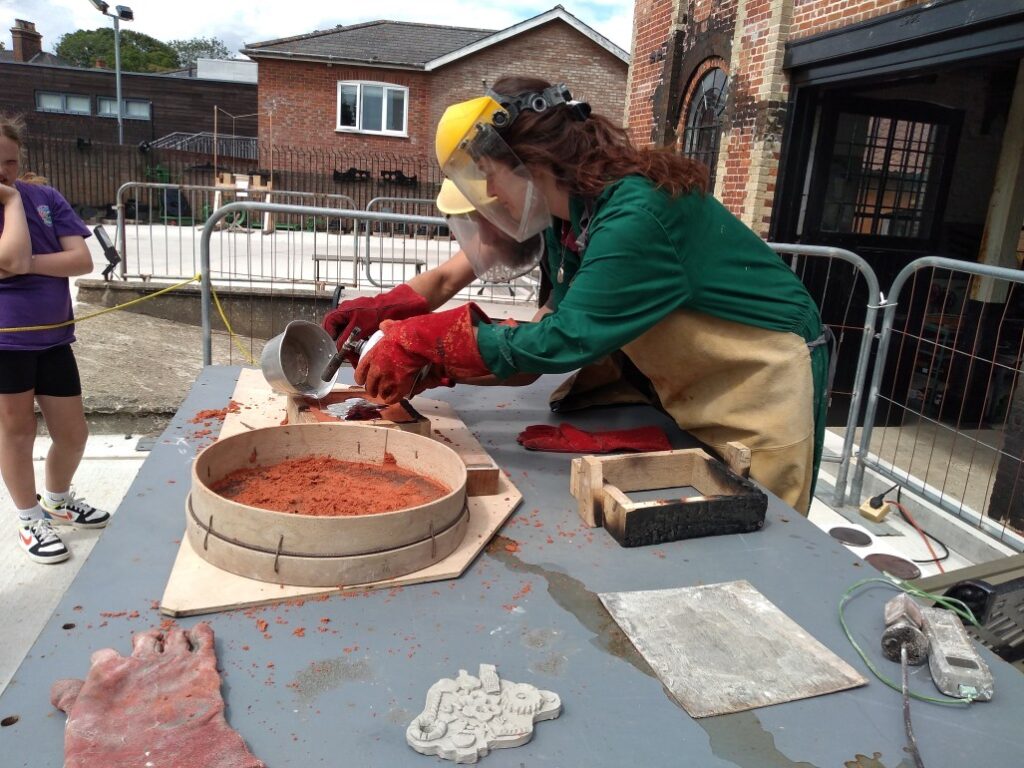 Sam and Ellie pour molten pewter into the shape left from the mould in the sand