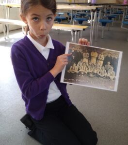 A school girl kneels on the floor, holding an old sepia photograph of a football team. She indicates on of the people in the picture and is pulling a long face, as if impersonating the individual