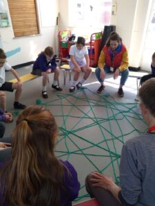 A group of children and adults seated on school benches. On the floor in front of them is a large 'net' of green wool, that they have created by throwing a ball of wool back and forth to one another