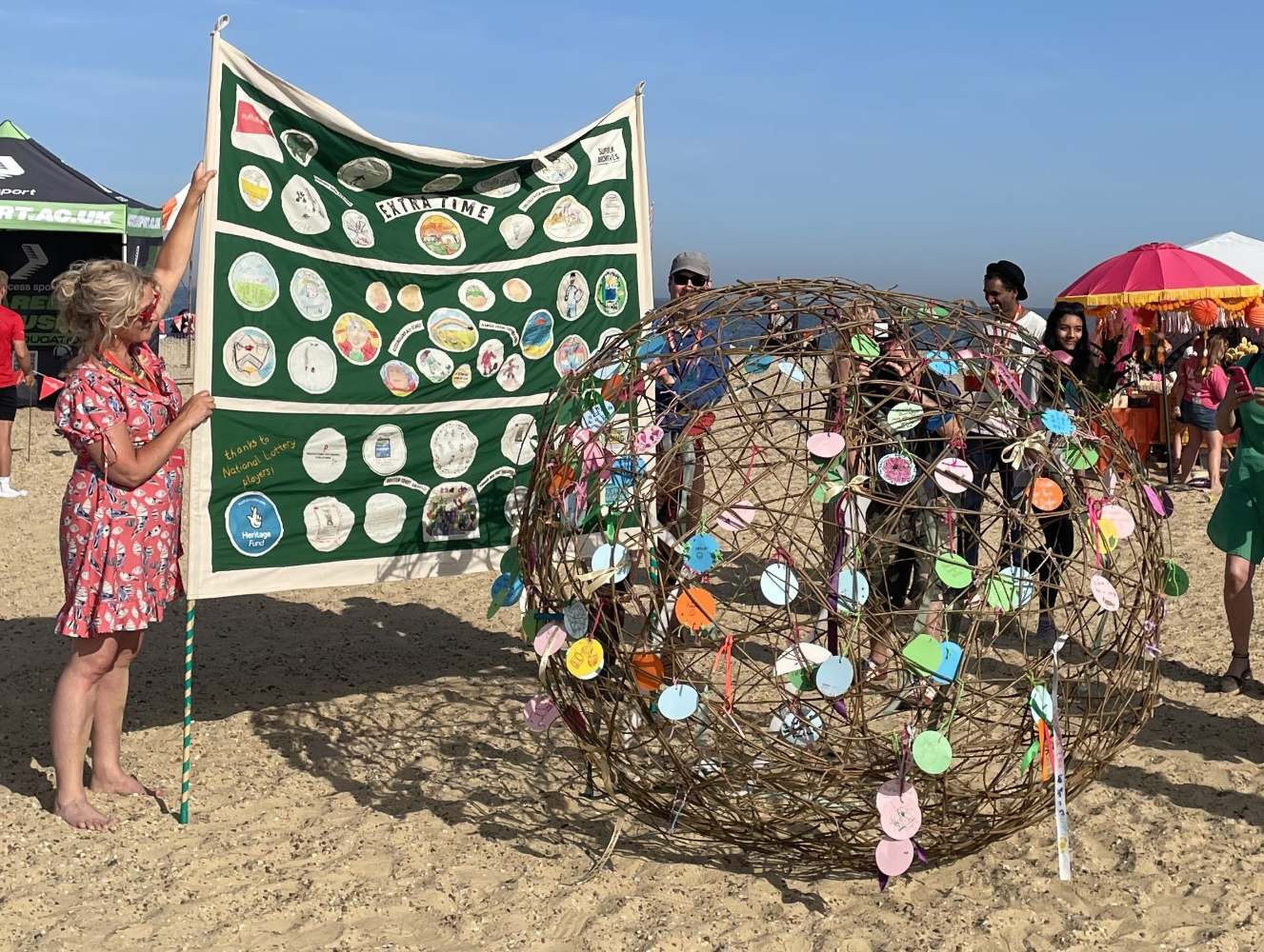 A woman stands on a sandy beach, holding up a large football banner decorated with colourful images and text. In the forefront is a large willow ball festooned with hand written 'goals' on card circles