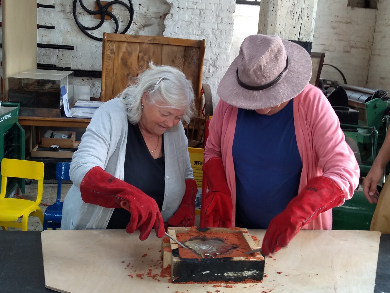Two adults, one wearing a large pink hat, are bending over a wooden mould with orange sand and a metal object in the middle. Each person wears a pair of enormous red heatproof gloves. They each have a long rod with which they are lifting the metal object out of the sand