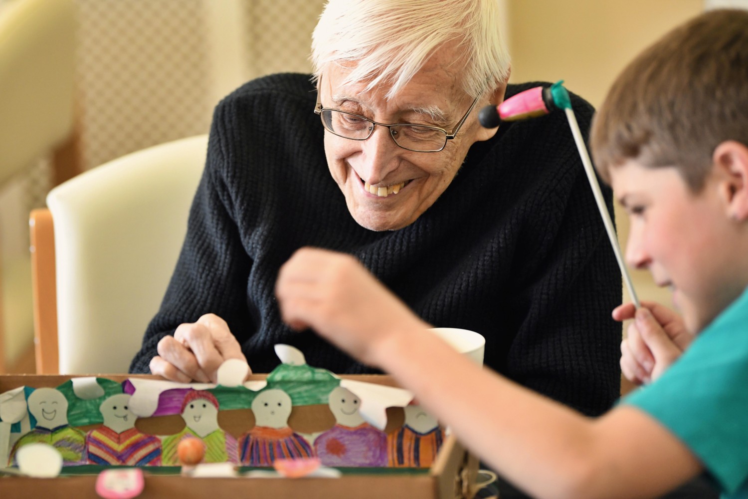 A gentleman wearing a dark jumper grins down at a cardboard football game whilst a child to his left is positioning something. The child is holding a long stick with a wooden figure taped to the end