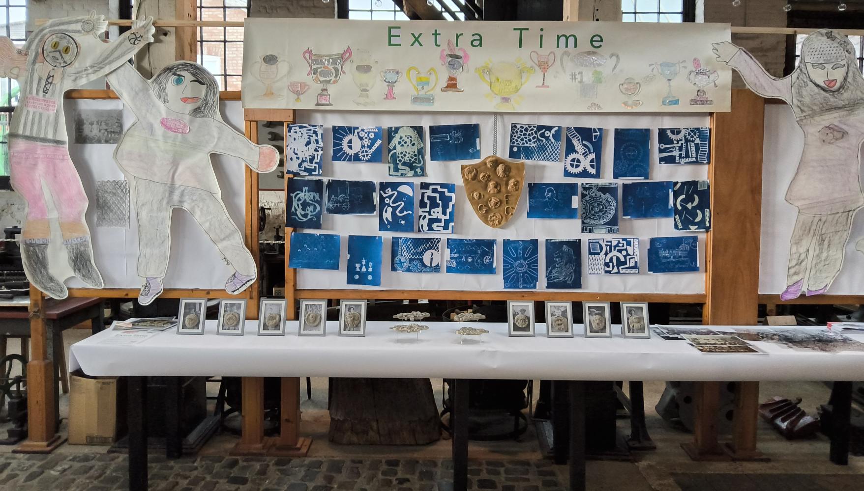 A display of cyanotypes flanked by life-sized football figures. Above is a large mural of handcrafted trophies and the title Extra Time and on the table in front is an arrange of photographs in grey frames and other objects
