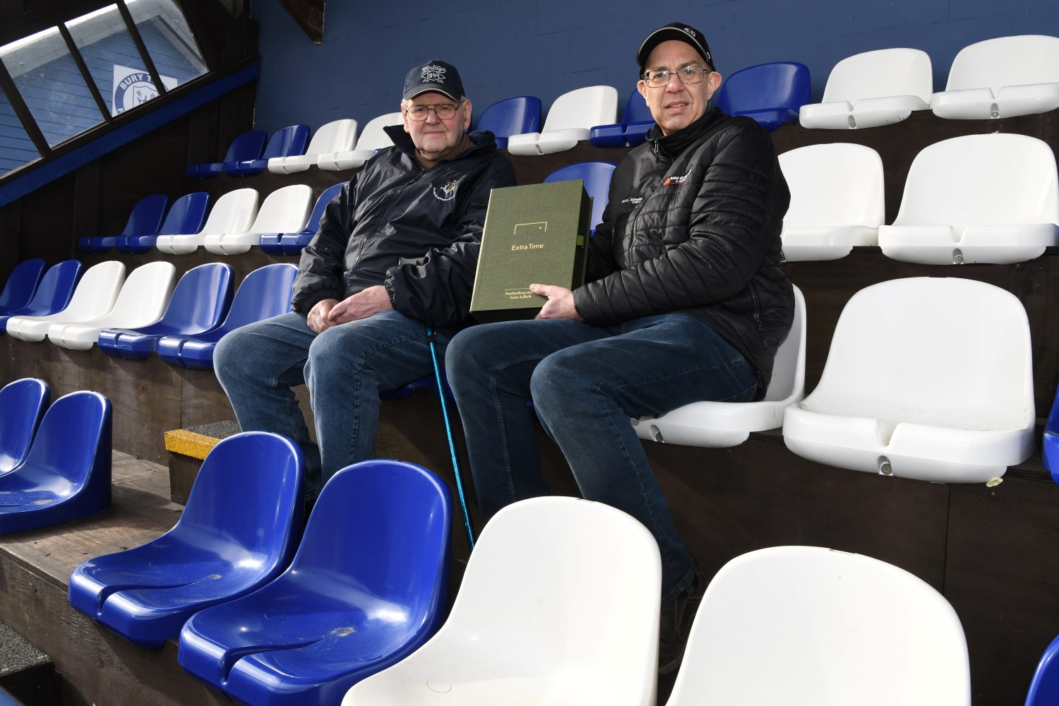Two gentlemen seated in blue and white seats at a football ground holding a green file box with Extra Time printed on the front