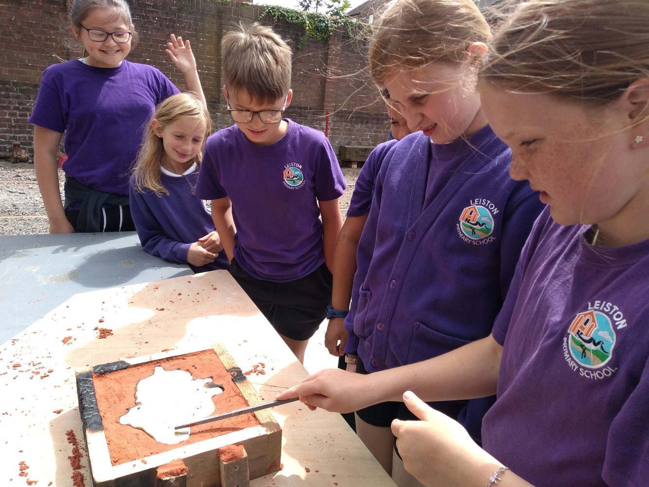 A line of school children in purple tops stand in front of a table on which is a wooden mould full of red sand with a metal object in the middle
