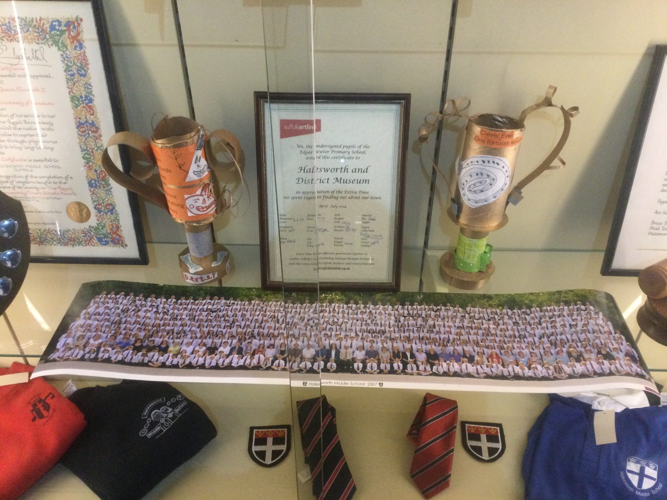 A glass cabinet full of school memorabilia. On bottom shelf are jumpers with school crest, 2 ties and 2 school badges. On the shelf above is a long photograph of all school pupils behind which stand 2 ornate cardboard trophies and a special certificate of attendance for Halesworth and District Museum