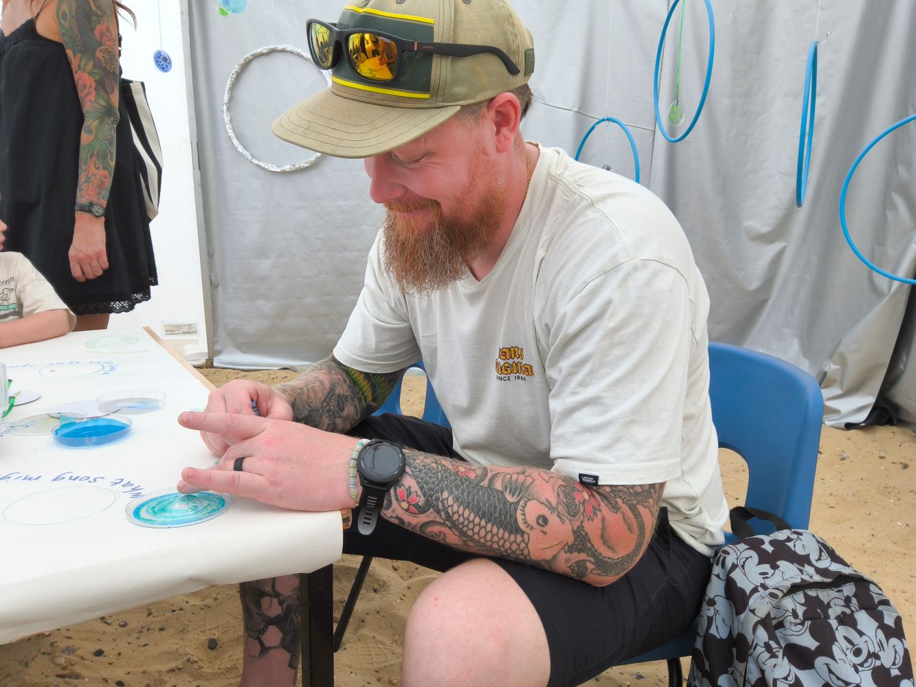A man seated at a table, with cap and sunglasses on head and tattooed arms, drawing a disc
