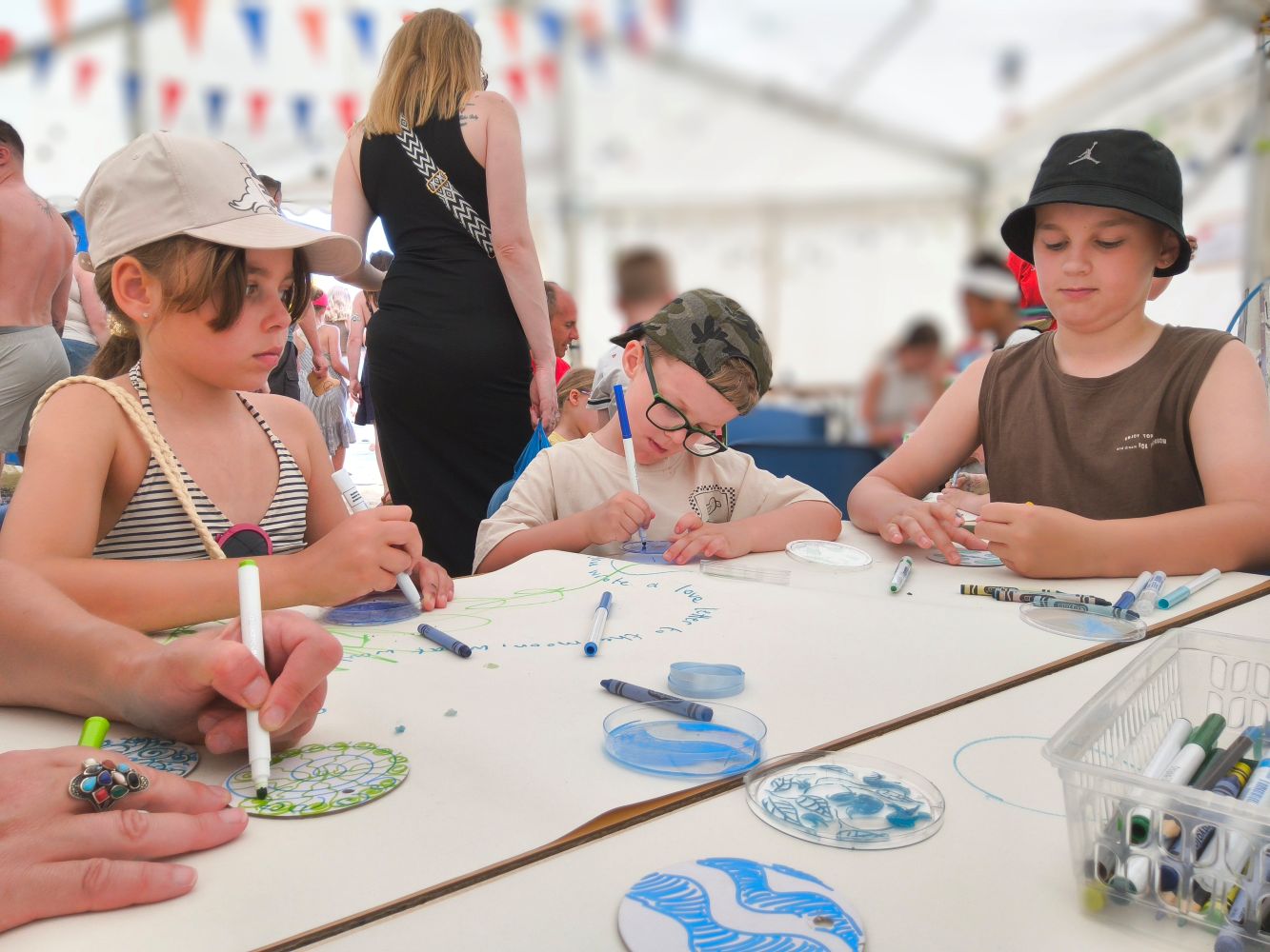 Three children seated at a table, adult in background, drawing on discs and Petri dishes