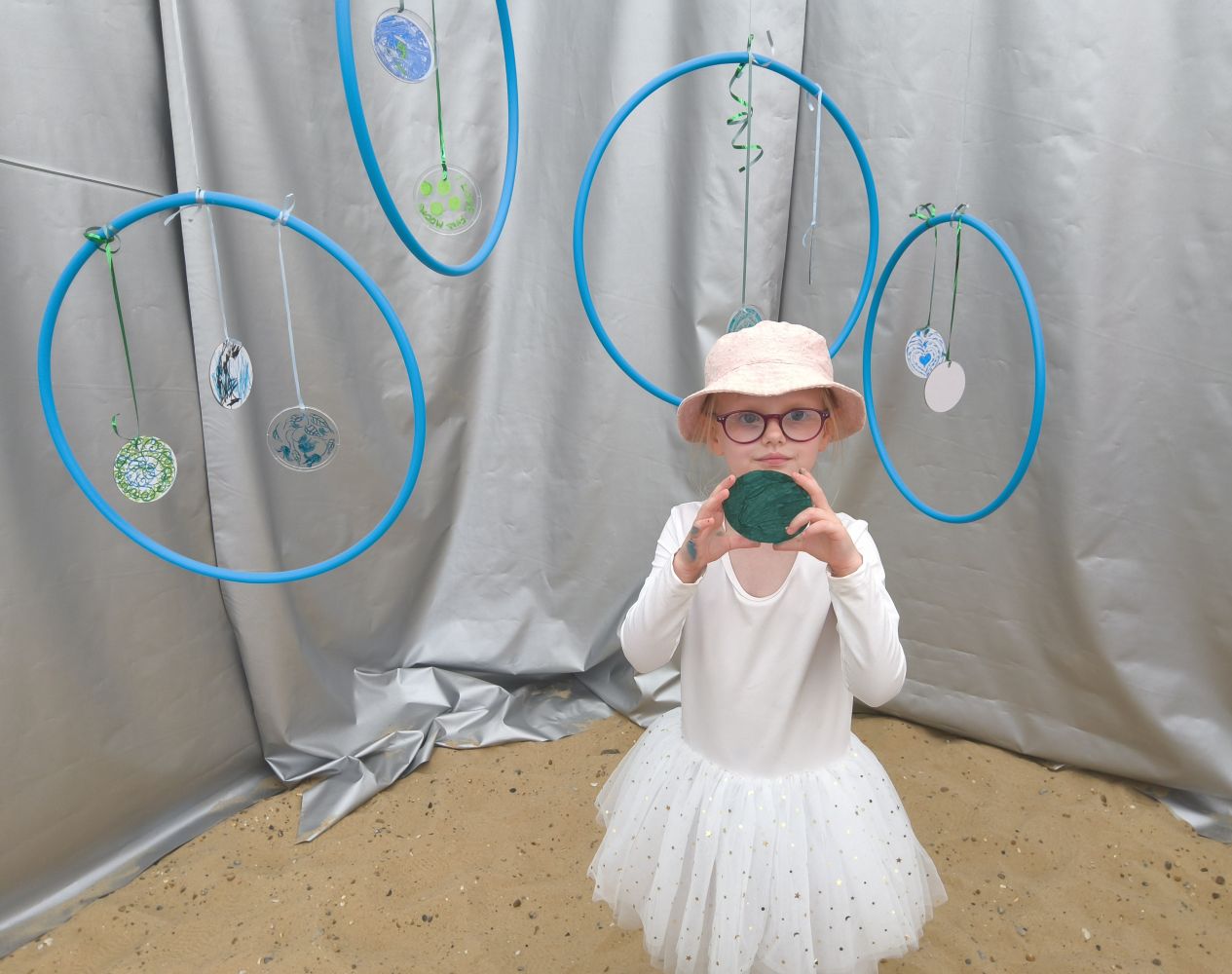 A child in white dress and sunhat stands in front of silver backdrop with blue hoops, she's holding a green moon she's made