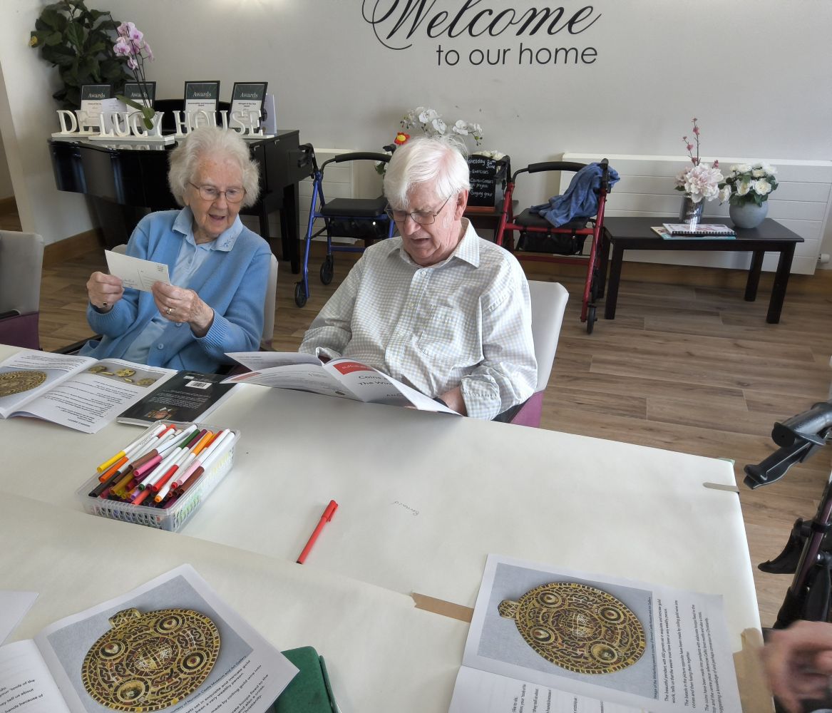 A man and woman seated at a table with magazines open and a basket of coloured felt tip pens