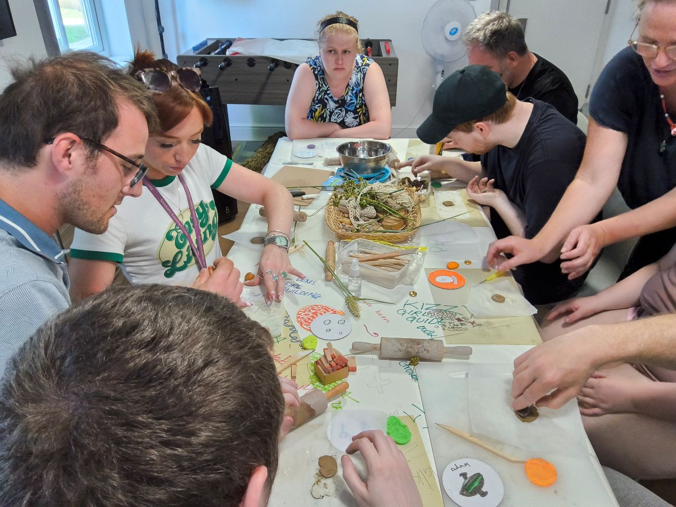 A crowded table, covered with rolling pins, plasticine and objects, with people busy rolling out and making
