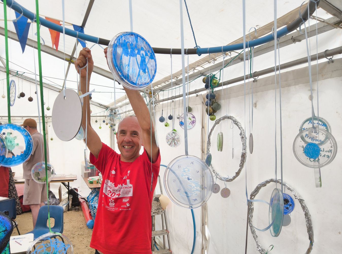 A man in a red T-shirt, grinning at camera as he adds his moon to a hoop of suspended discs