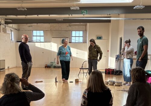 A cropped shot where 8 musicians in a circle are visible. They are in a hall with some standing and some sitting.