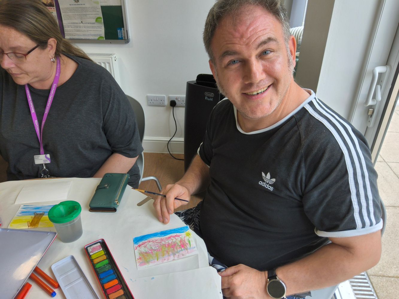 A person grins at camera. On the table in front of him is a paint box and other painting materials. A person with a purple lanyard is seated on his right