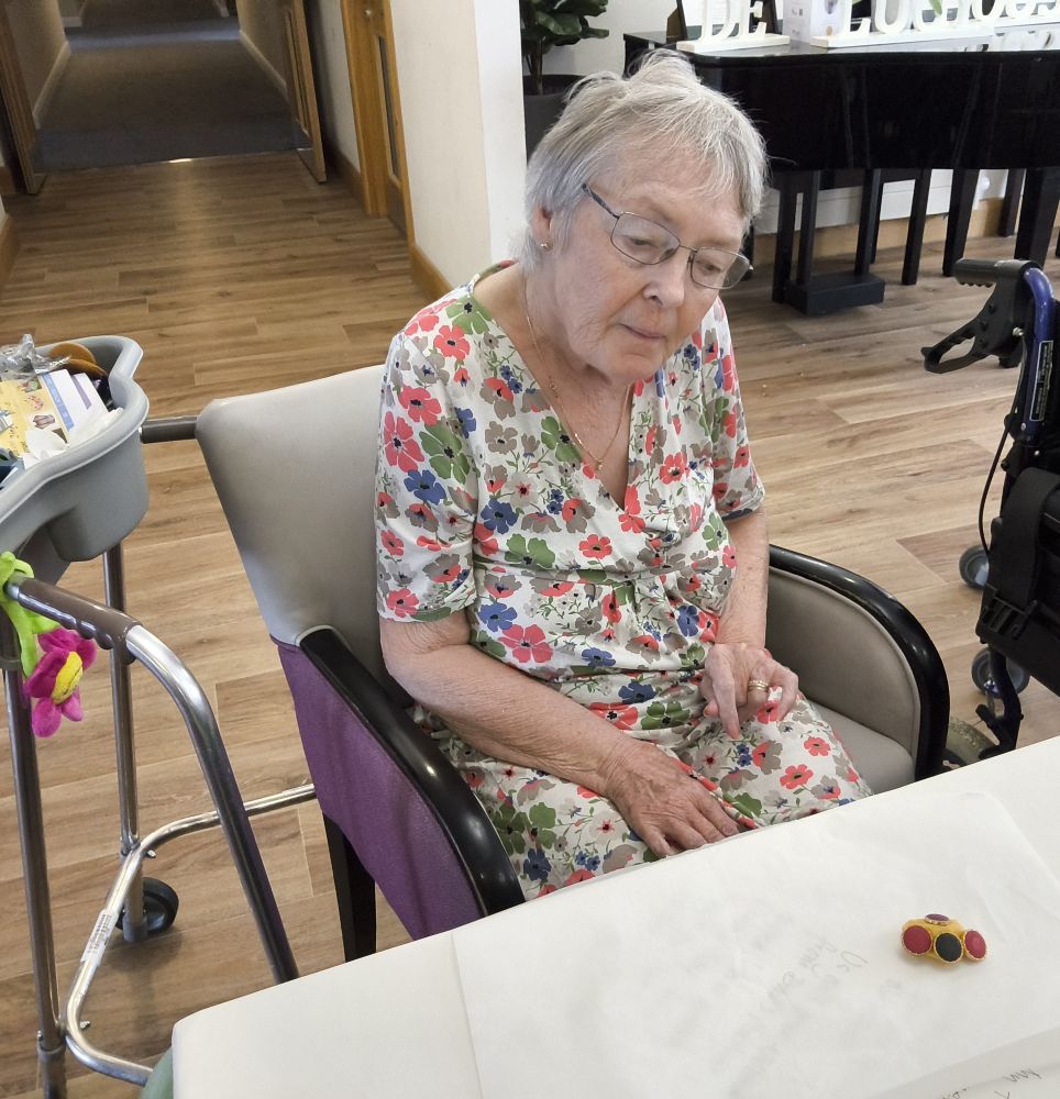 A woman in a floral frock is seated by a table on which is a small object covered with small red and black buttons