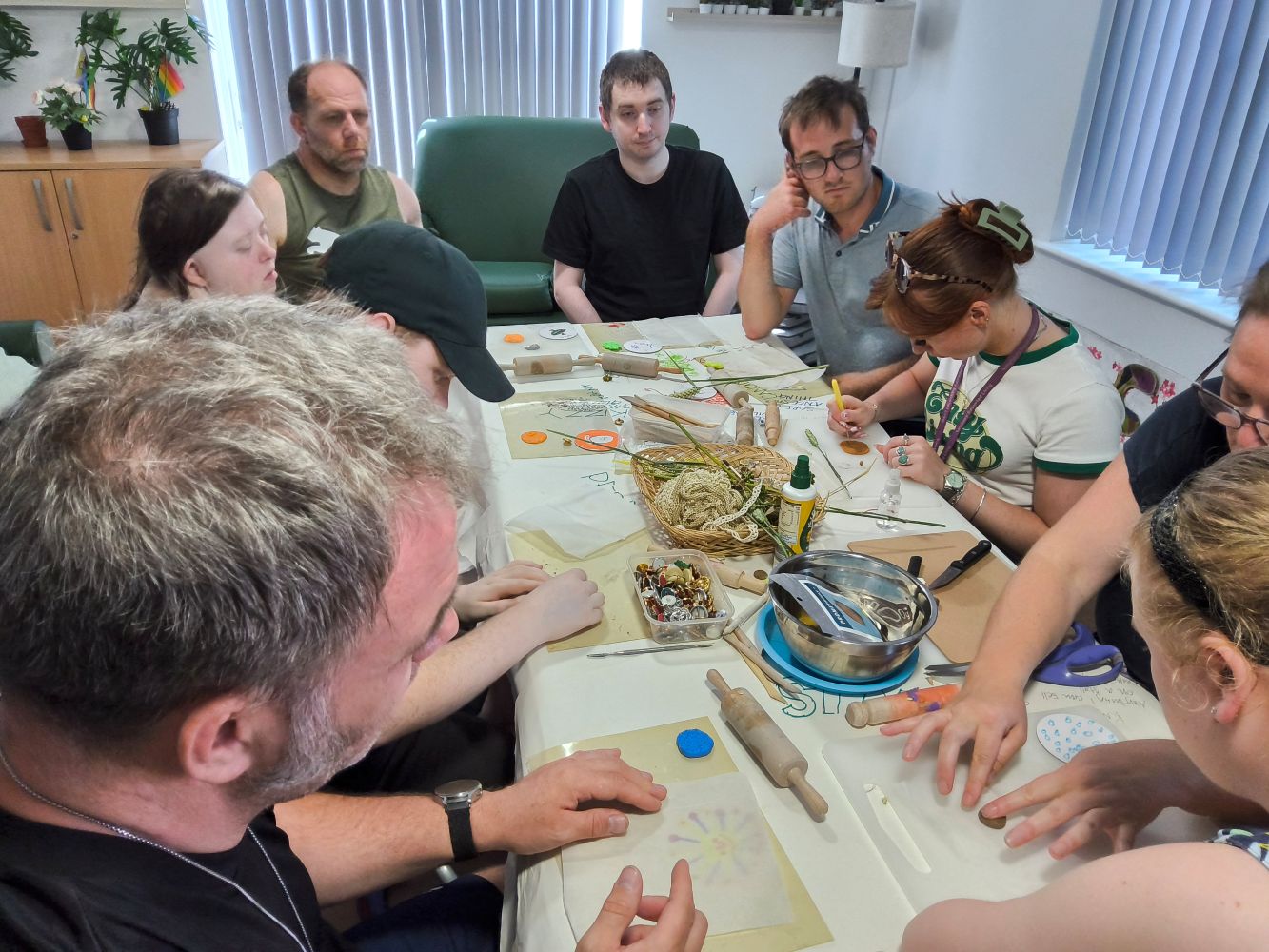 Several people seated around a table, some watching a others roll out and shape clay