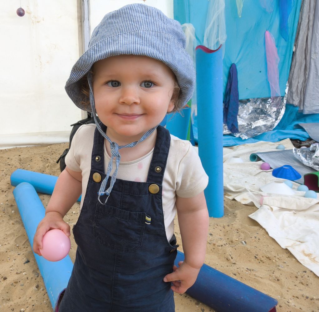 A toddler in blue dungarees and sun hat, holding pink ball in right hand, smiles at camera