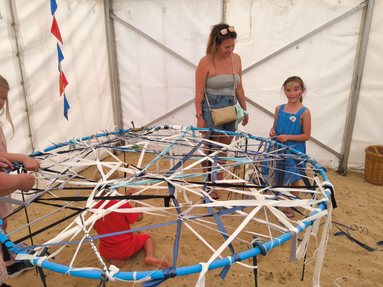 A mother and daughter working at the giant weaving hoop. A child in a red top is underneath, threading a ribbon through it