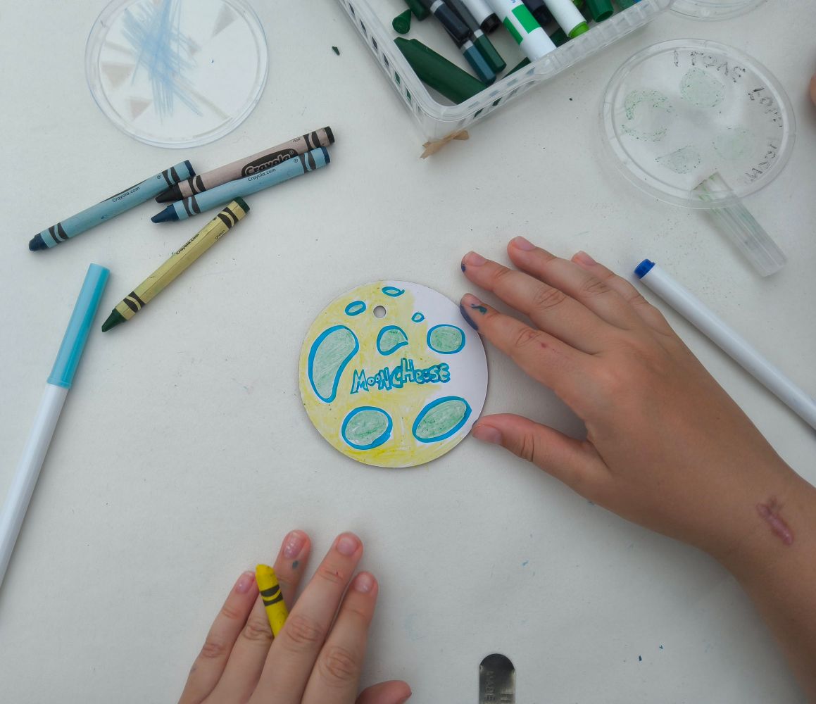 A coloured disc with the word moon cheese surrounded by crayons