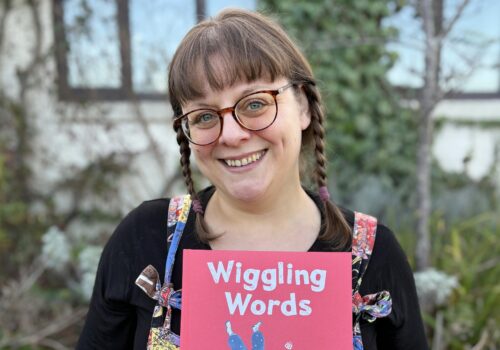 lady with brown braided hair and glasses holding a children's book with a red front cover. standing outside a building with foliage in background.