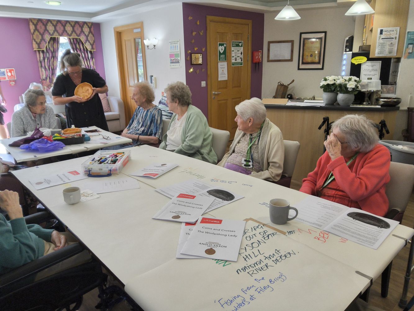 A woman dressed in black and holding a gold-coloured bowl is standing at the end of a long row of tables around with are seated several people. On the table are some publications, mugs and art materials. In the background is a small kitchen area with vases of artificial white flowers on the counter