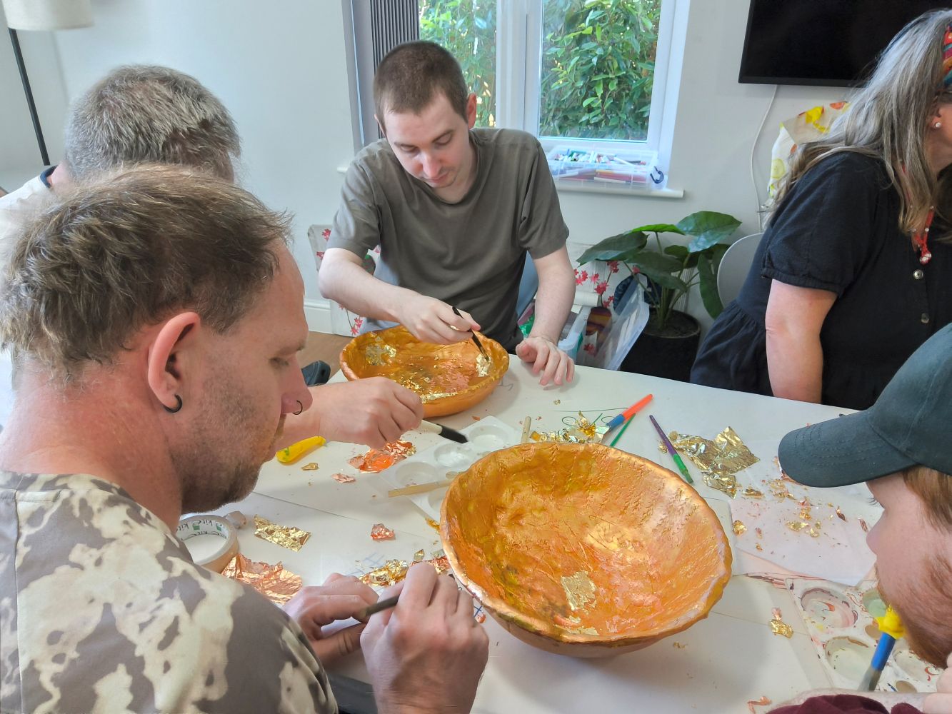A group of four young men applying gold leaf to two gold bowls, with lots of bits of leaf scattered around the table