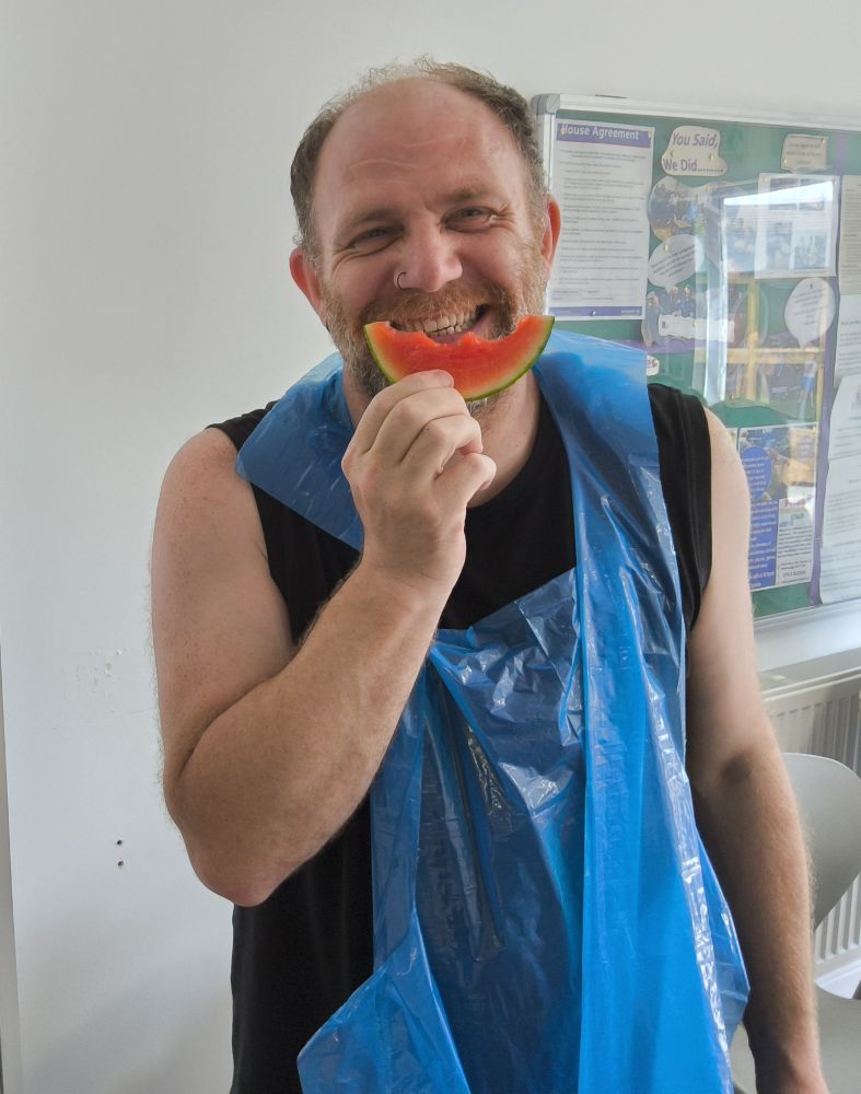A man in a black sleeveless T-shirt and a blue plastic apron smiles at camera, holding a slice of watermelon in front of his face