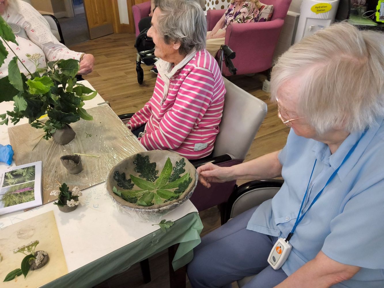 Two women seated at a table, one in a pink and white striped top and the other in a short sleeved blue blouse. She is touching the rim of a grey clay bowl, inlaid with leaves, and on the table is more clay and bunches of greenery