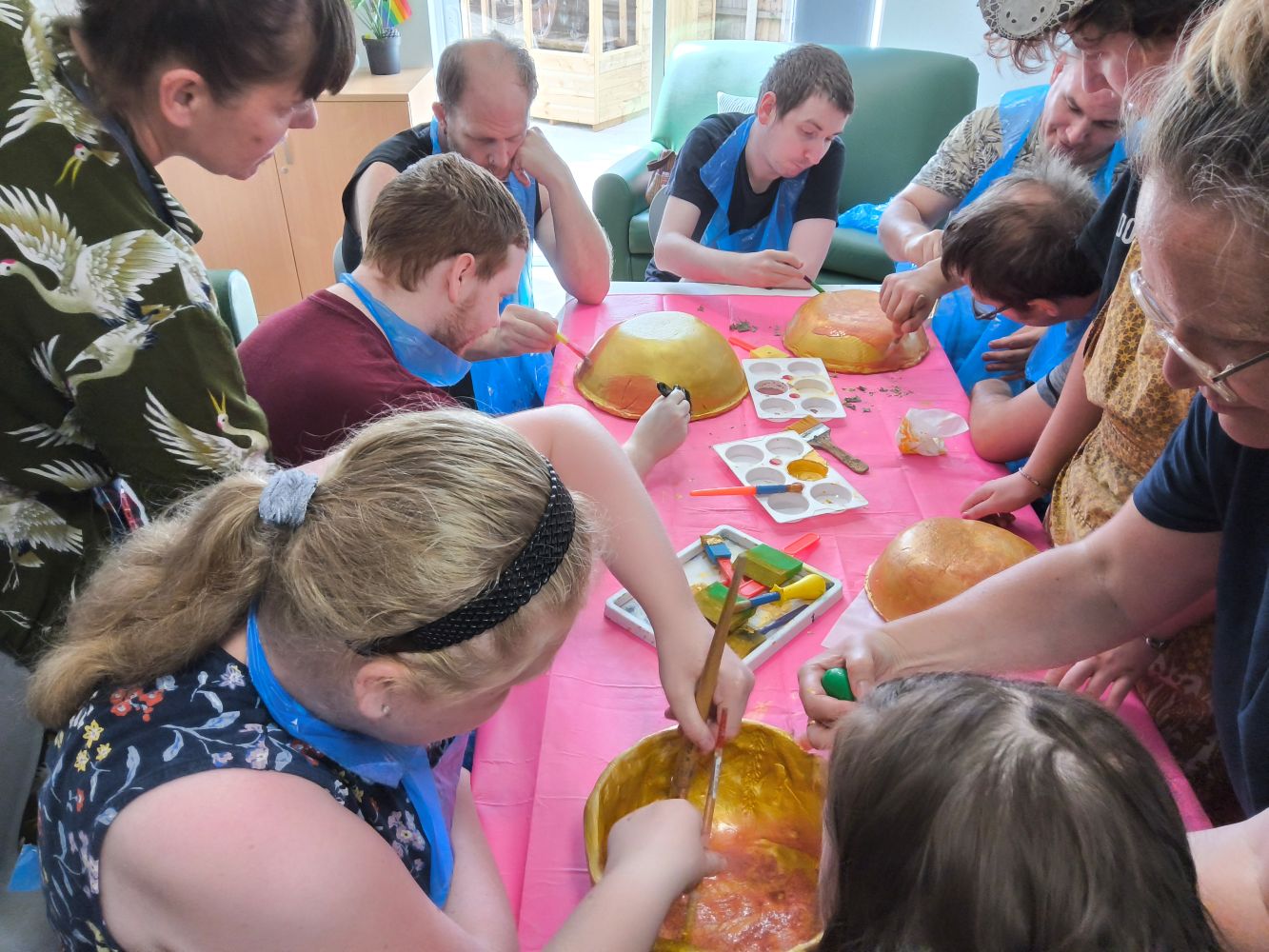 A group of people around a table covered in a pink cloth. Some are seated, some standing. They are all applying gold paint to bowls