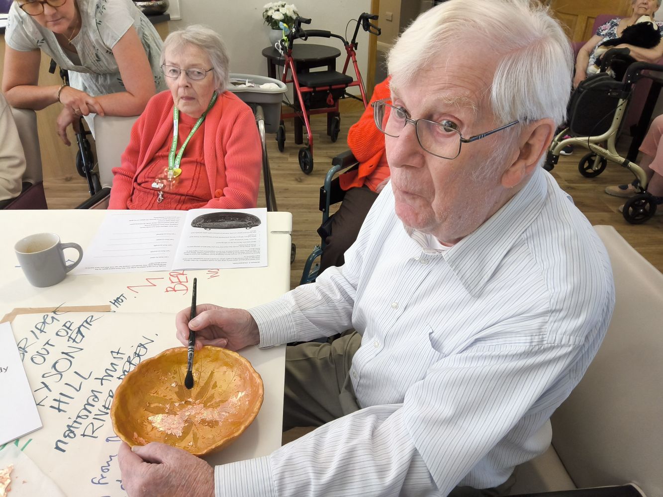 A gentleman seated at a table, wearing spectacles and a striped open neck shirt. He is brushing gold leaf into a bowl. He is watched by a woman in an orange top and wearing a green lanyard