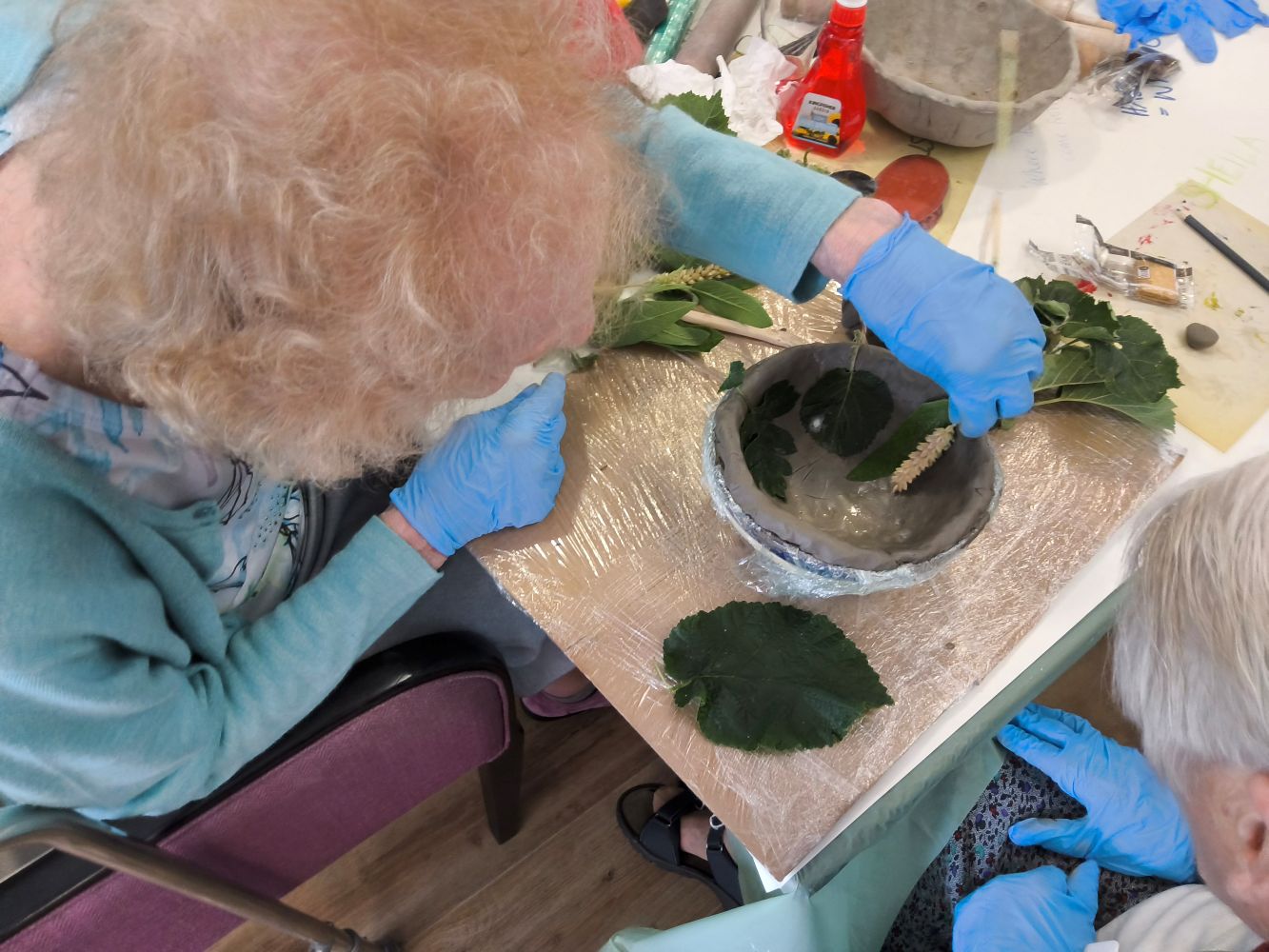 A woman wearing blue gloves leans over a clay bowl, lining it with leaves and an ear of wheat