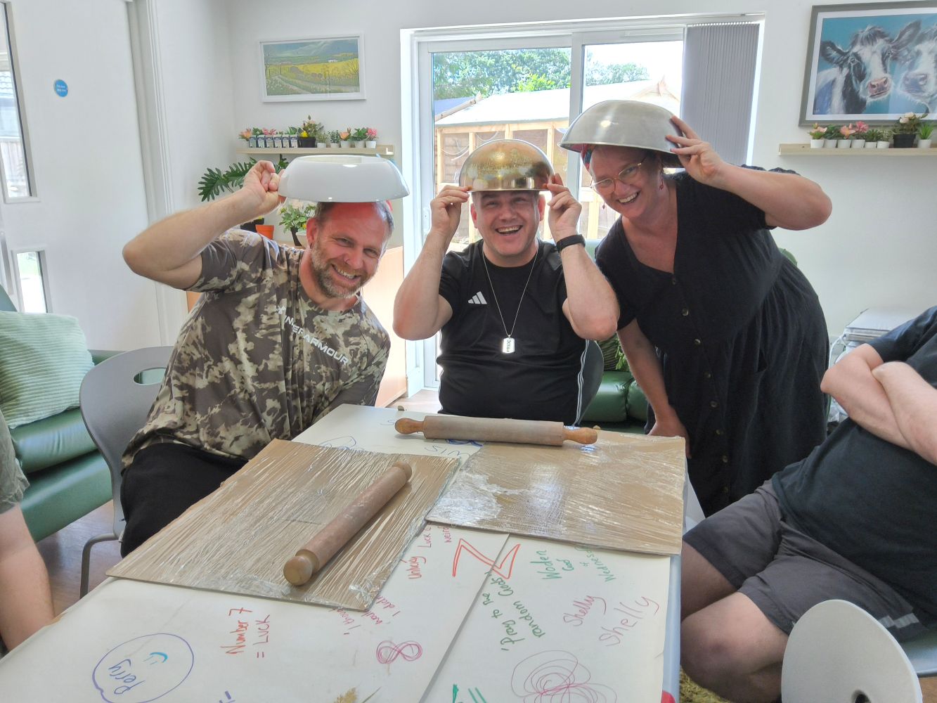 Three people pose for the camera, balancing bowls on their heads. On the table in front are two board covered with cling film and a pair of large wooden rolling pins