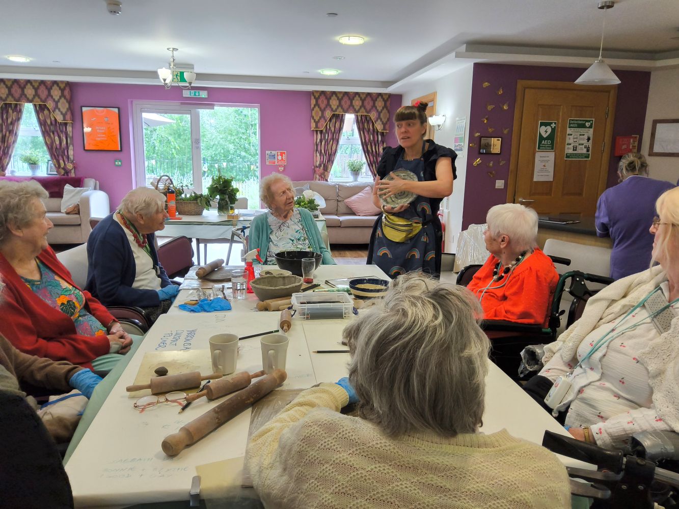 A woman stands, holding a bowl in her hands. In front is a large table with elderly ladies seated all around. On the table is an assortment of mugs, rolling pins and art materials