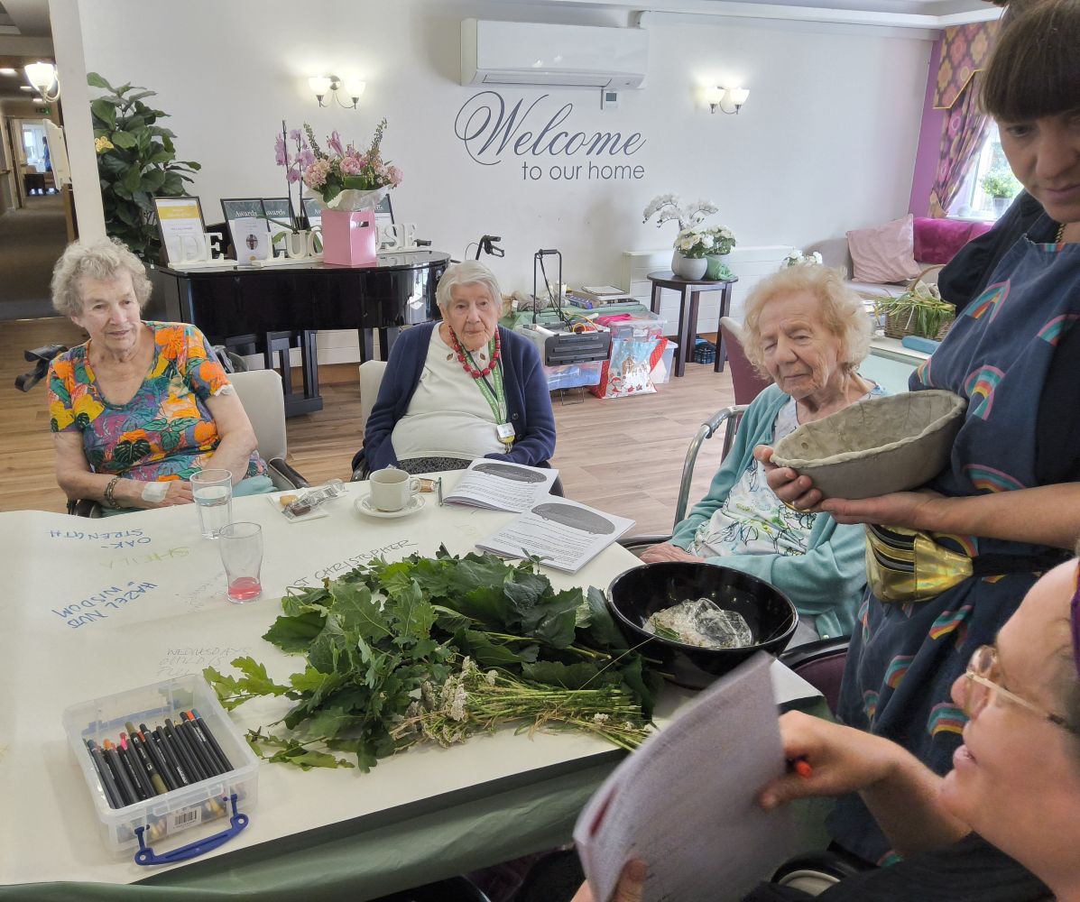 Women seated around a table on which is a large bunch of greenery, a box of pencils and some magazines opened at a picture of a bowl. A woman standing is holding a clay bowl.