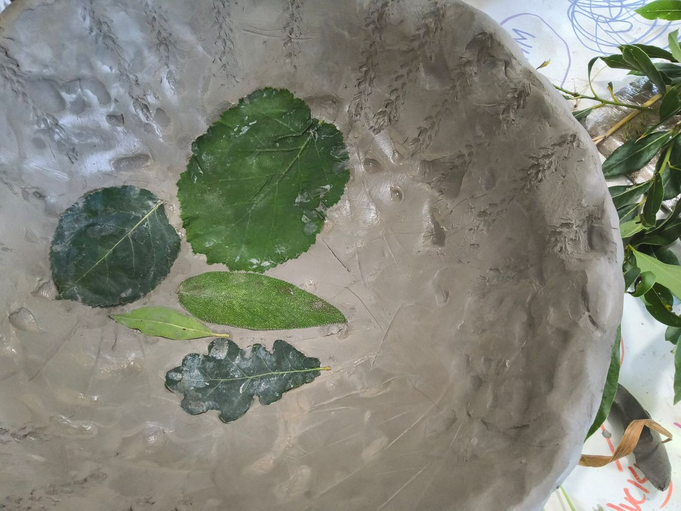 A wet clay bowl with leaves stuck in the bottom and a pattern around the top made from an ear of corn