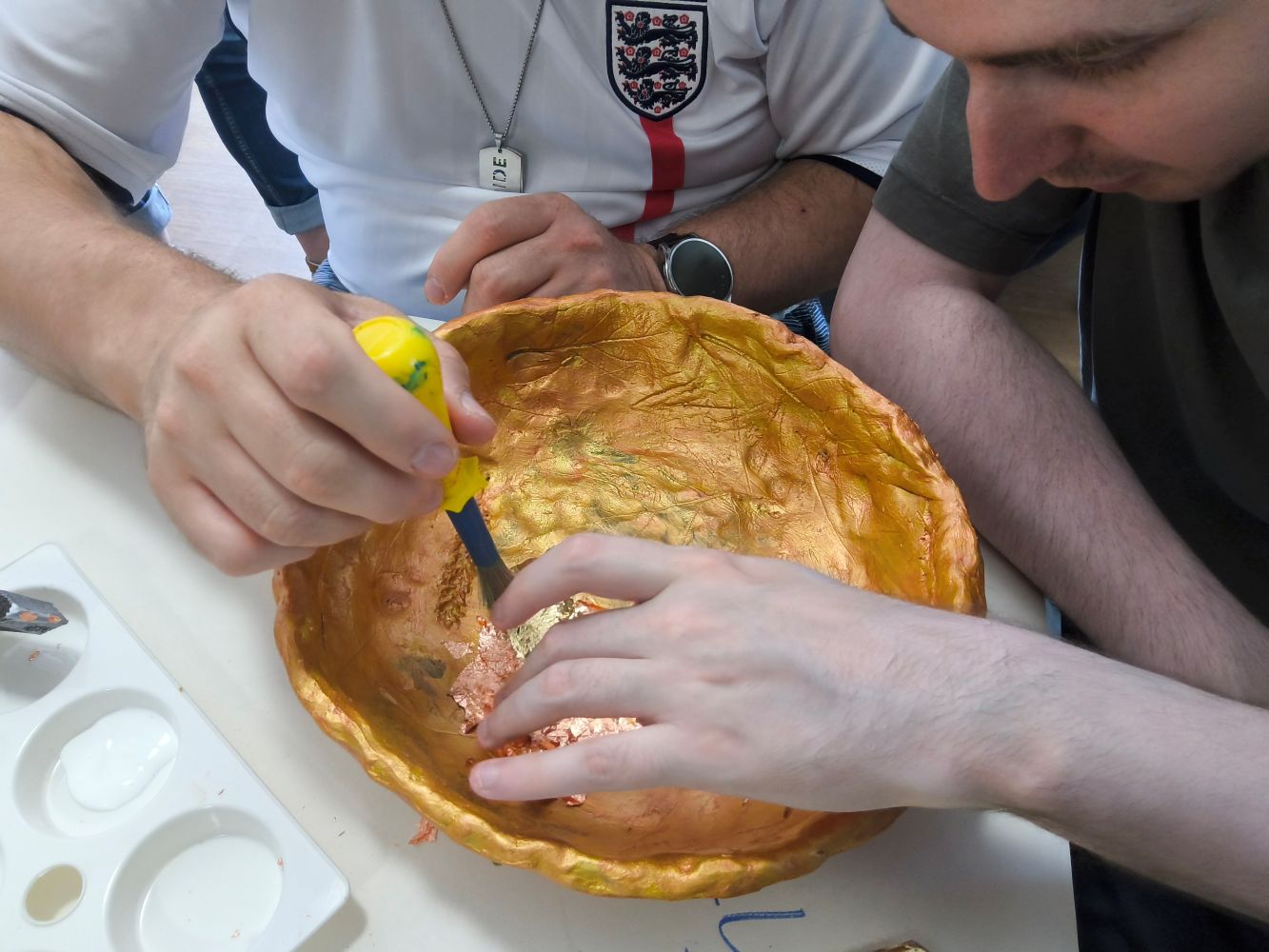 Figures bent over a bowl, one holding a yellow-handled brush whilst the other uses his finger to gently apply the gold leaf