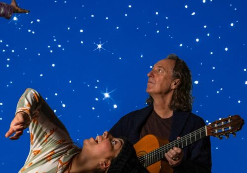 2 performers looking up with a star filled background. 1 performer has a guitar.