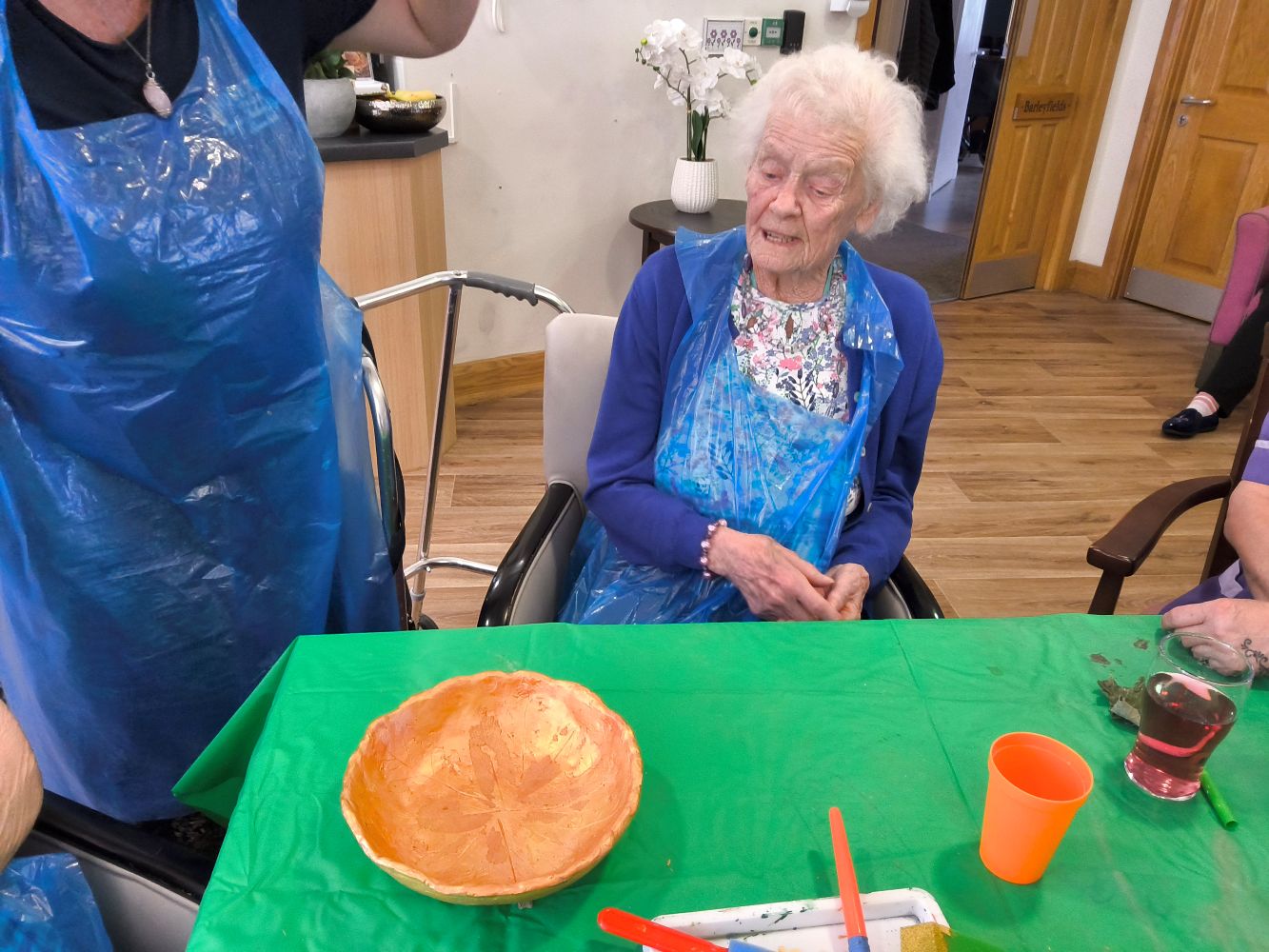 A woman seated, wearing a floral top, blue cardigan and blue plastic apron, admiring a gold-painted bowl on the table in front of her
