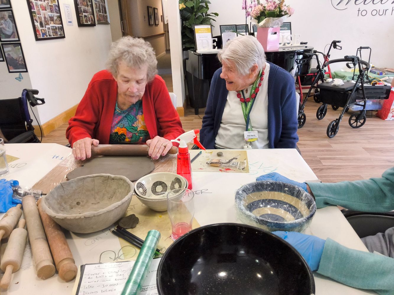 A woman wearing a floral top and red cardigan is rolling out a piece of clay in front of her. Seated beside her is a lady wearing a white top, blue cardigan and several beads and lanyards round her neck. On the table is an assortment of bowls and rolling pins