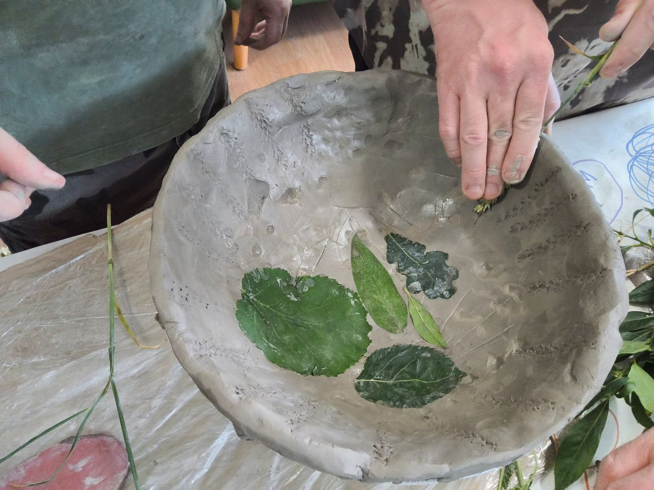 Some leaves in the base of a wet clay bowl and a hand using a piece of wheat to make a pattern around the rim